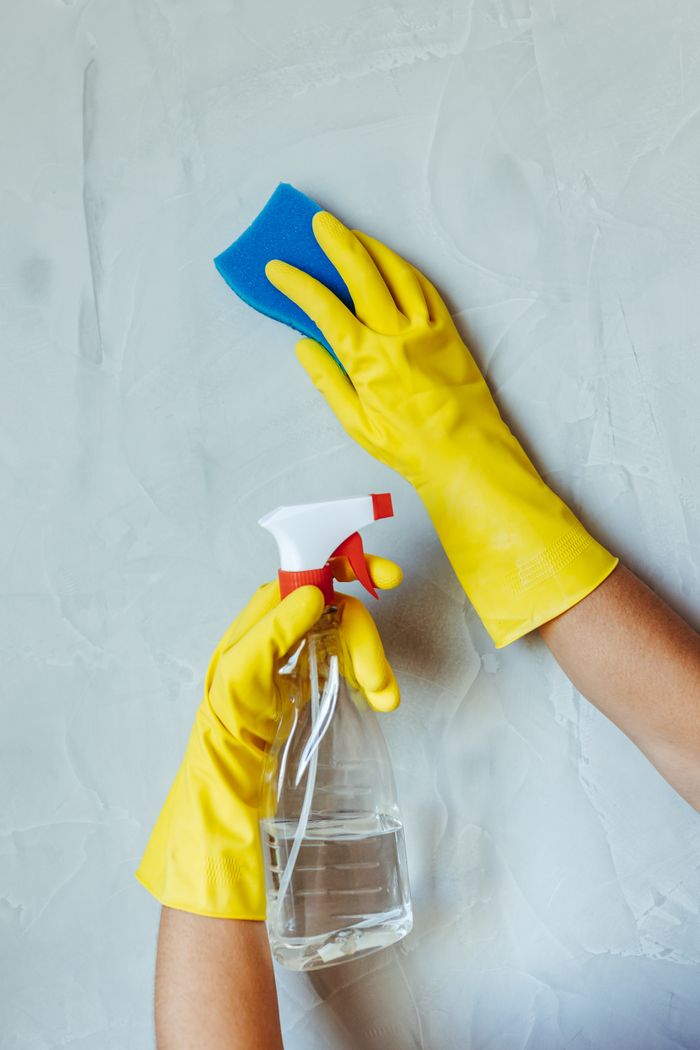 Yellow-gloved hands cleaning a wall with a sponge and spray bottle.